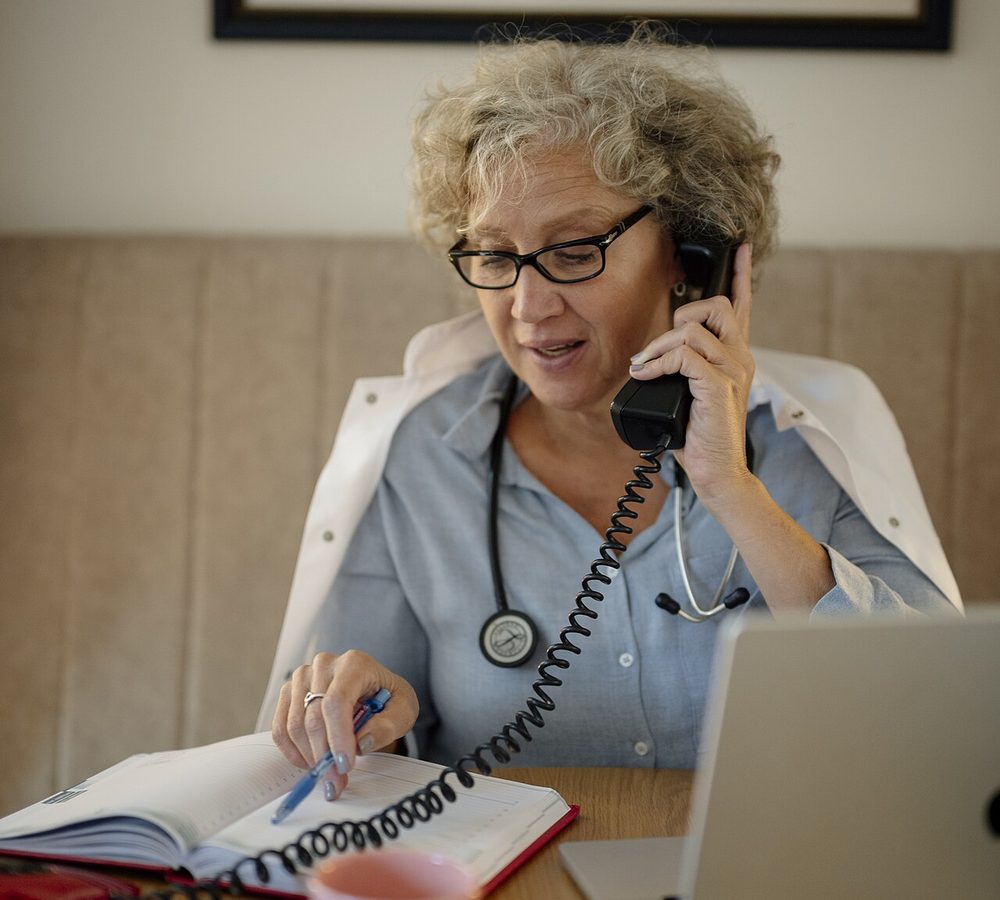 A doctor with a stethoscope talking with a patient on the phone while taking notes at her desk